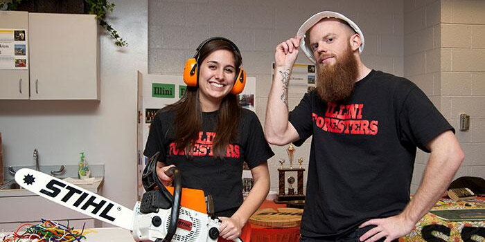 Two students with safety gear and a chainsaw